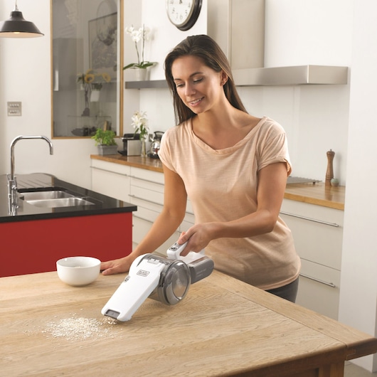 A person is using a BLACK+DECKER handheld vacuum cleaner (model PV1820L) to clean spilled oats from a wooden kitchen table. The kitchen is modern with a red island, white cabinets, and a sink in the background. The person’s face is obscured.