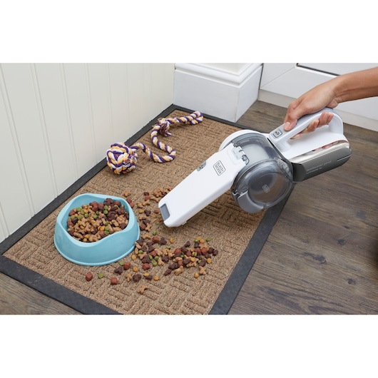 A hand using a BLACK+DECKER handheld vacuum cleaner to pick up spilled pet food from a mat next to a pet bowl and a rope toy.