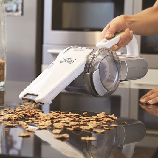 A person using a BLACK+DECKER handheld vacuum cleaner to pick up cereal flakes from a countertop in a kitchen.