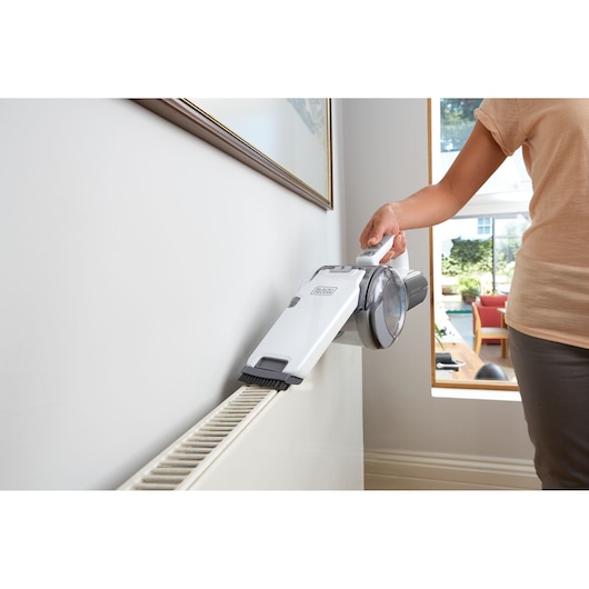 A person uses a BLACK+DECKER PV1820L handheld vacuum cleaner to clean the surface of a radiator in a bright home interior.