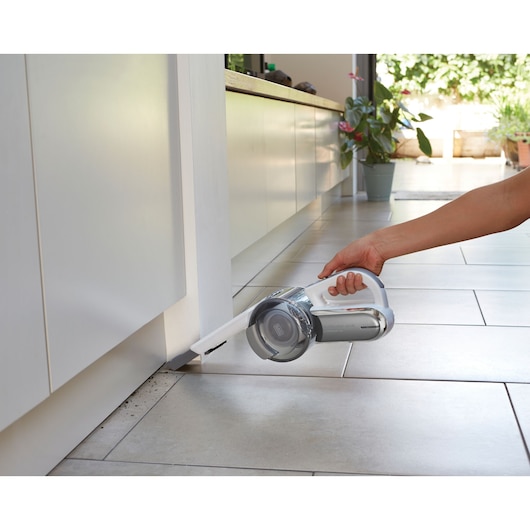 A person using a BLACK+DECKER handheld vacuum cleaner with a narrow nozzle attachment to clean dust from the edge of a tiled floor near a kitchen cabinet.