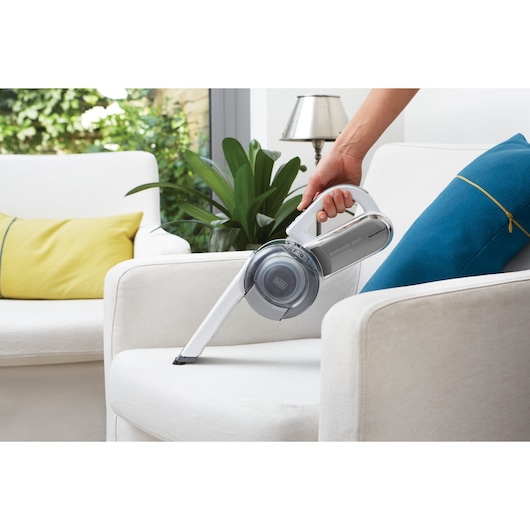 A close-up of a person using a BLACK+DECKER handheld vacuum cleaner to clean a white armchair in a modern living room. There are decorative pillows and a green plant in the background.