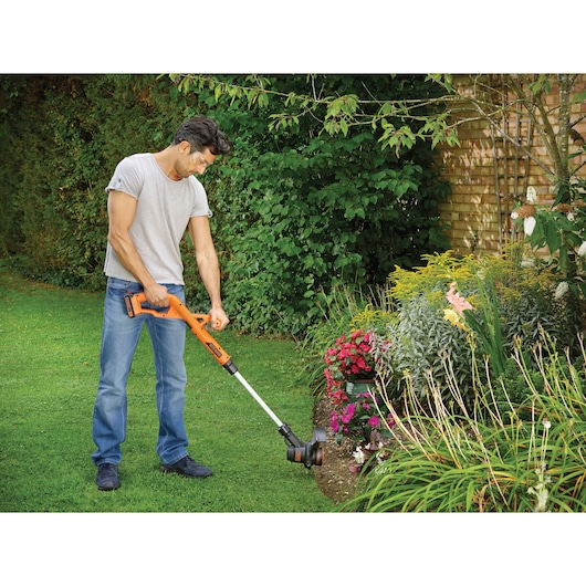 A person using an orange and black BLACK+DECKER cordless grass trimmer to edge a garden lawn next to flower beds in a backyard.