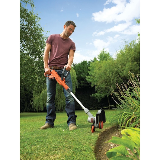 A person is using a BLACK+DECKER STC1820 cordless grass trimmer to edge a lawn in a garden. The trimmer is orange and grey with a handle and wheels. The garden has green grass, various plants, and trees, with a partly cloudy sky in the background.