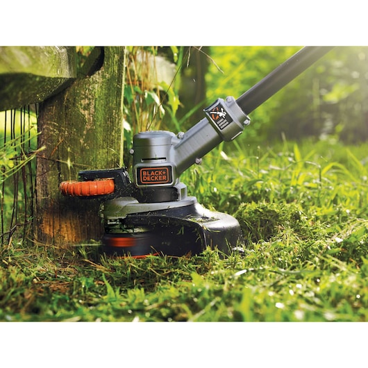 Close-up of a BLACK+DECKER cordless grass trimmer in use, cutting grass along the edge of a wooden fence outdoors.