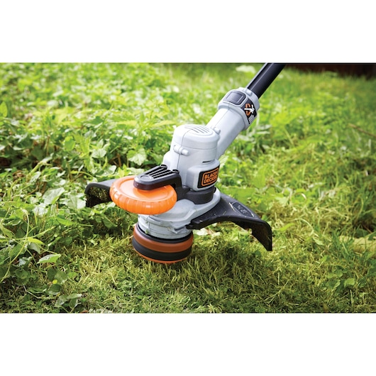 Close-up of a BLACK+DECKER cordless grass trimmer in use, trimming green grass and weeds outdoors.