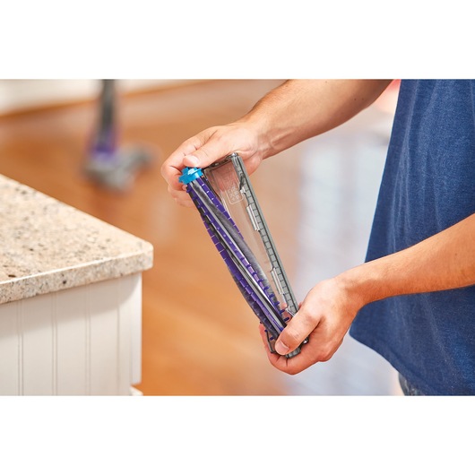 A person holding a vacuum cleaner brush roll attachment, with a focus on the hands and the device, indoors on a wooden floor near a countertop.